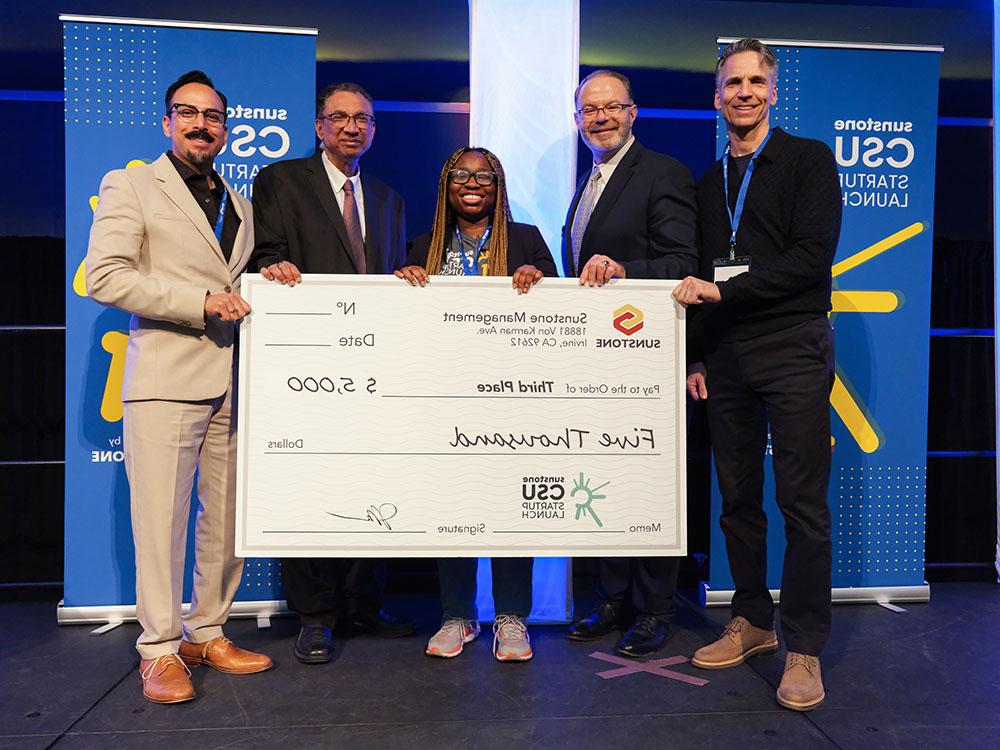 A female, college-age startup founder poses onstage with two sponsors, two hosts, and a giant check for $5,000