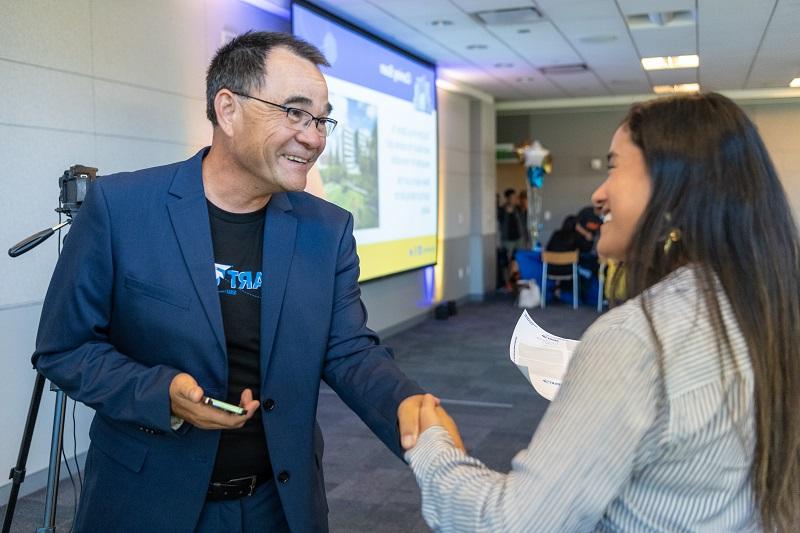 A woman in a striped blouse with long, dark hair shakes hands with a smiling man with short, salt-and-pepper hair.  He wears a black t-shirt with the SpartUp logo under a blue suit. 