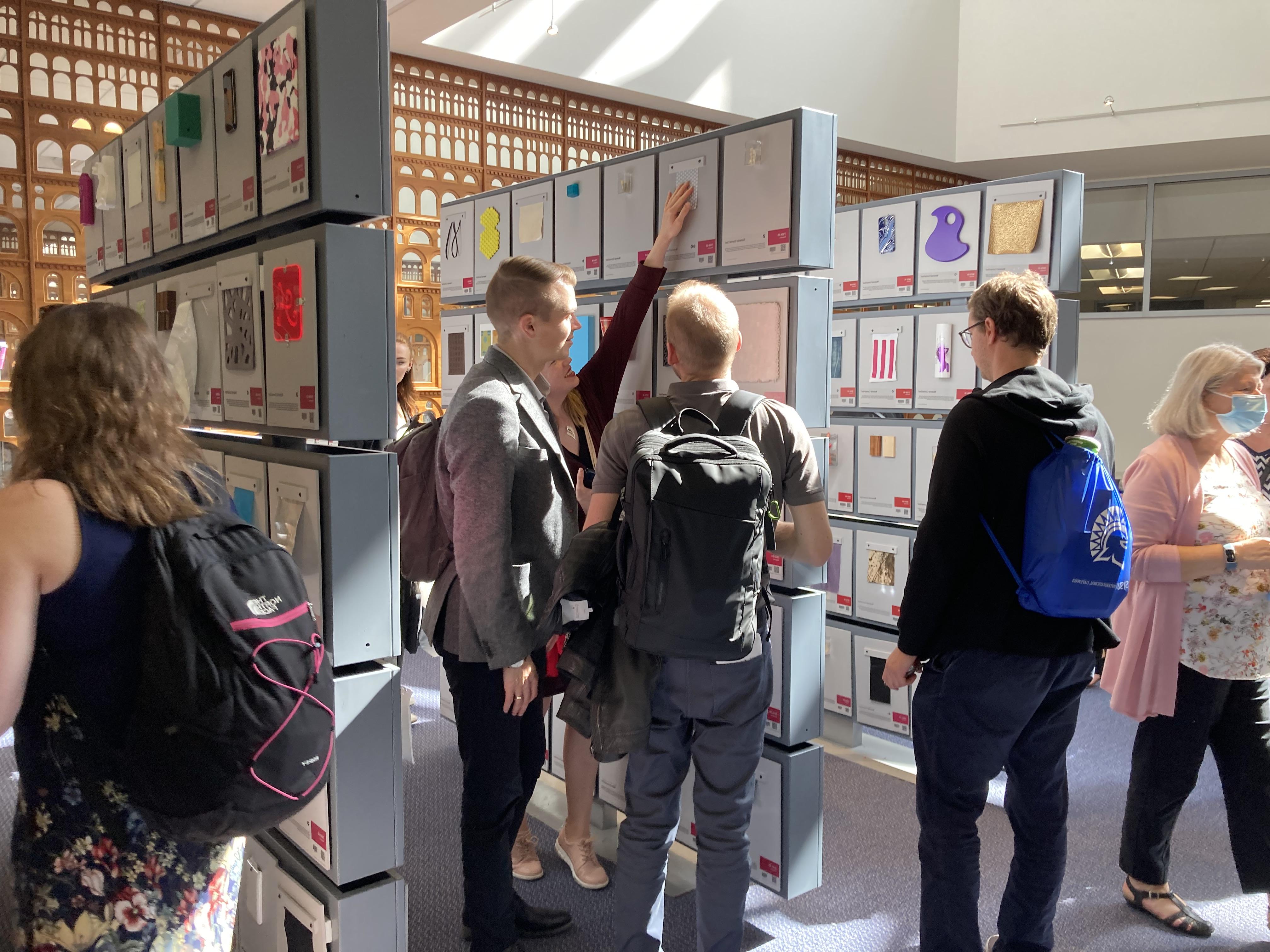 Three male students and one female stand in front of a panel of squares, each with a 4' scrap of plastic, fabric, or other material in 菠菜网lol正规平台's Materials Library.  A staff member with gray hair and a pink sweater walks away in the background, while a woman with a background walks to the right in the foreground.